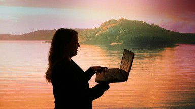 The profile of a woman is seen using a laptop against a picturesque background of a lake with land in the distance 