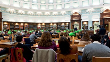 Suíonn cuairteoirí sa Seomra Léitheoireachta i Leabharlann Náisiúnta na hÉireann, ag amharc ar bhean ag labhairt le linn imeacht Poetry Aloud 