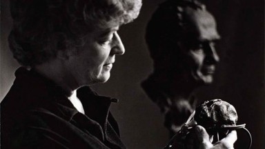 Grianghraf dubh agus bán de Helen Hooker O'Malley le píosa dealbhóireachta ina lámh, agus dealbh bhrád sa chúlra