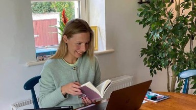 Tugann bean spléachadh ar leabhar agus í ina suí os comhair a ríomhaire glúine oscailte