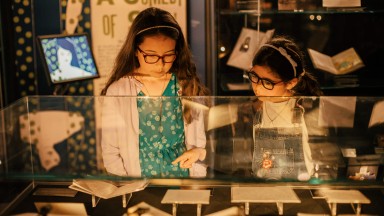 Two young girls wearing glasses examine exhibition materials in a glass case