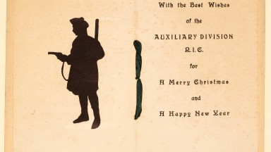 A yellowed greeting card bound by ribbon shows a silhouette of a soldier pointing a pistol, with text on the page opposite that reads: "With the best wishes of the Auxiliary Division R.I.C. for a Merry Christmas and a Happy New Year" "Ireland, Christmas, 1920"