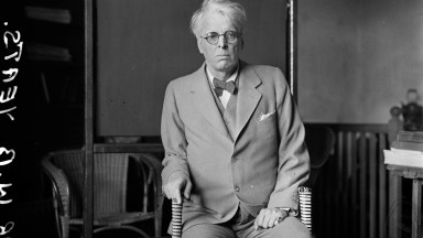 WB Yeats ag caitheamh culaithe, ina shuí i gcathaoir agus ag baint beagáinín taca as bata siúil 