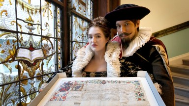 Two actors in period costume pose with grant of arms next to stained glass window