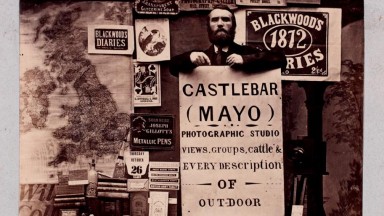 Sepia photograph of a man holding a large sign advertising his Photographic Studio in Castlebar, Co.Mayo