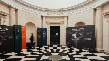 Rotunda in Georgian style of architecture. The floor has alternating black and white tiles. Standing in front of the walls are signs with images of Seamus Heaney and quotes from his poetry. The is a person dressed in black walking around the space on the left. They are blurred and their face is not shown.