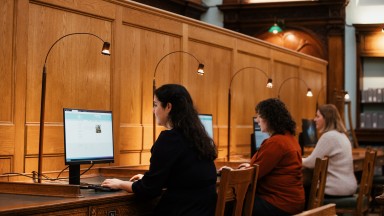 Suíonn triúr ban ag déanamh taighde ar scáileáin ríomhaire taobh istigh de phríomhsheomra léitheoireachta an NLI