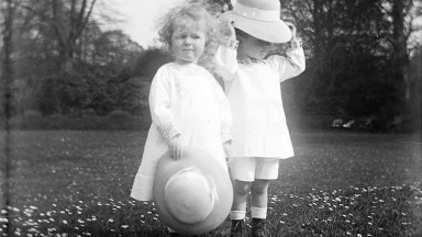 Two toddlers with bonnets standing in field surrounded by daisies