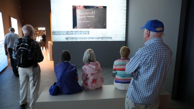 Photograph of a dozen people watching a large screen within the Seamus Heaney: Listen Now Again exhibition. 