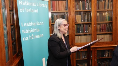 Woman speaking from a podium with bookshelves in the background