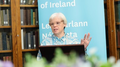A woman giving a speech in front on a blue banner and bookshelves