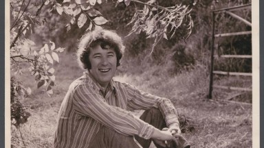 Black and white photograph of man, Seamus Heaney, sitting on grass in a striped shirt.