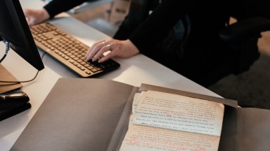 Image of hands typing at a computer with manuscript on desk