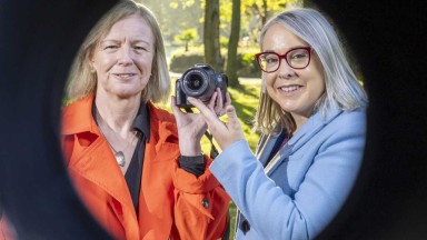 Two women holding a camera pointed at the viewer, with black circular frame 