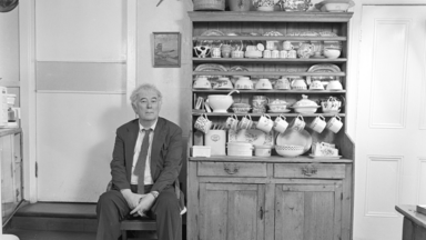 Seamus Heaney sitting in a wooden chair in a rural kitchen next to a cabinet filled with crockery