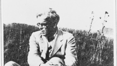 Black and white photograph of poet W. B. Yeats sitting in a grassy field. He is dressed in a light-colored suit and glasses, with his arms wrapped around one knee, gazing to his left. The setting is natural and serene, with tall grass and wild plants in the background.