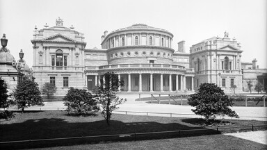 Black-and-white photograph of the National Library of Ireland in Dublin, showing its grand neoclassical facade. The central rotunda with tall columns and circular windows is flanked by two ornate rectangular wings topped with statues. In the foreground, landscaped gardens with trees and pathways frame the library building.