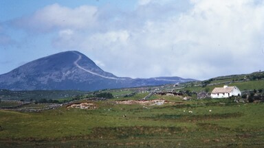 A scenic landscape featuring a large, rounded mountain with a visible trail leading to its summit, surrounded by green fields and stone walls. In the foreground, a white cottage with a thatched roof sits among rolling grassy hills under a partly cloudy sky.