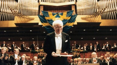 seamus heaney in formal evening attire stands on stage holding a certificate and medal during an award ceremony. Behind him, a large audience applauds, and a blue and yellow banner with the year “1901–1995” hangs above a grand pipe organ. The setting is ornate and ceremonial, with a medallion relief visible on a podium nearby.