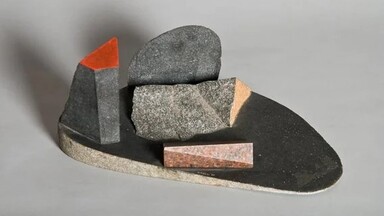 bstract stone sculpture composed of geometric and organic forms in shades of gray, black, and reddish-brown. The arrangement includes angular blocks and curved pieces set on an oval base, with one element featuring a bright red triangular top, evoking a minimalist landscape or architectural composition.