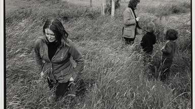 Black-and-white photograph of a young Marie Heaney walking through tall grass in a rural field, looking down as wind bends the grass around her. In the background, Seamus Heaney stands near a fence with two young children, all partially obscured by the grass, with open farmland stretching behind them.