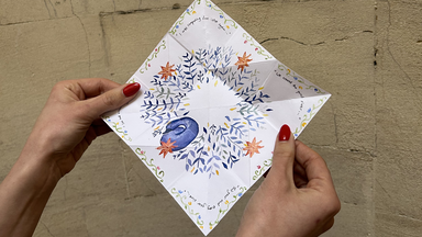 hands hold the paper fully unfolded into a square, showing a detailed hand-painted design of blue leaves, small orange flowers, forming the feathers of a blue peacock at the center, with handwritten poetry along the edges.