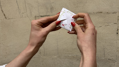 ands with red nail polish hold a small, neatly folded piece of illustrated paper against a beige stone wall.
