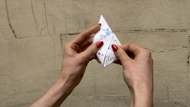 Two hands with red nail polish hold a small, neatly folded piece of illustrated paper against a beige stone wall. Only a triangular corner of the artwork is visible.