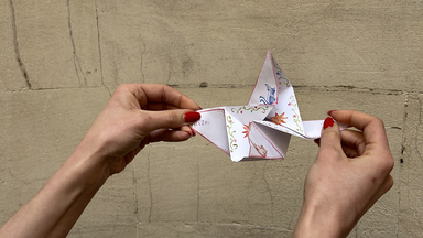 hands gently pull the folded paper open, revealing a zigzag of unfolding panels with delicate floral illustrations on white paper.
