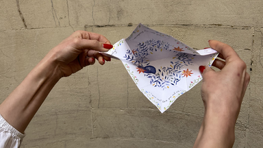 hands gently pull the folded paper open, revealing a zigzag of unfolding panels with delicate floral illustrations on white paper.