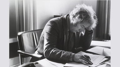 A black and white photo of Heaney writing poetry at his desk, holding his head in his hand