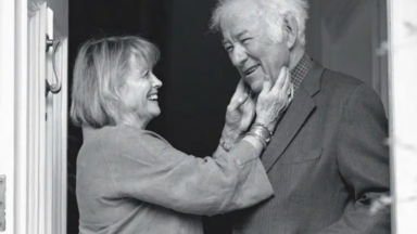 Black-and-white photo of Marie Heaney standing in a doorway, smiling warmly as she gently holds Seamus Heaney's face. Heaney, wearing a suit jacket, smiles back with one hand in his pocket, sharing an affectionate, intimate moment.