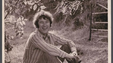 Black-and-white photograph of a young Seamus Heaney sitting on grass in a rural field, smiling at the camera. He wears a striped long-sleeve shirt, trousers, and lace-up boots, with his arms loosely wrapped around his knees. Tree branches with leaves hang above him, and a wooden gate stands in the background. 