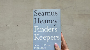 Hand holding a light blue copy of Finders Keepers by Seamus Heaney against a curved stone wall background.