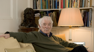 Seamus Heaney sitting on a yellow sofa in his home in front of a bookcase