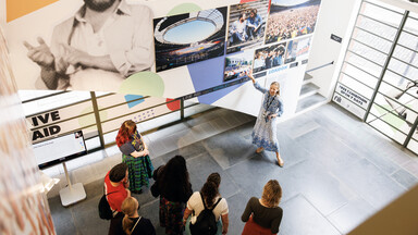 A museum guide gestures toward a large exhibition wall while leading a group of visitors through a gallery.