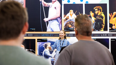 A female tour guide smiles and gestures while speaking to a small group of visitors in front of a large exhibition wall featuring iconic concert photographs. A prominent image of Freddie Mercury, shirtless and performing with a microphone dominates the display, with additional photos of other musicians arranged around it. The visitors are seen from behind, focused on the guide during the tour.
