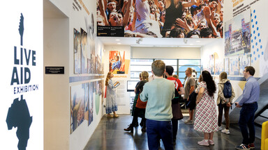 A museum guide leads a group of visitors through an exhibition featuring large concert photographs and displays.