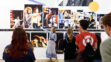 A museum guide points to a wall of concert photographs while leading a group of visitors through an exhibition.