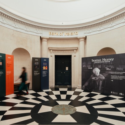 Rotunda in Georgian style of architecture. The floor has alternating black and white tiles. Standing in front of the walls are signs with images of Seamus Heaney and quotes from his poetry. The is a person dressed in black walking around the space on the left. They are blurred and their face is not shown.