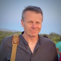 Alan Gilsenan standing outdoors in soft evening light, wearing a dark shirt and carrying a tan shoulder strap bag. He has short hair and a calm, thoughtful expression, with greenery and a pale blue sky in the background.