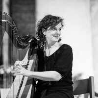 Black-and-white photo of a woman seated and playing a large harp, smiling and looking to her right, with soft focus background.