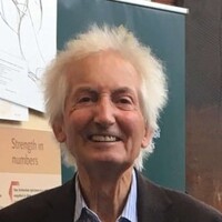 Image of Dr Cusack smiling warmly at the camera. He is wearing a dark blazer over a light blue striped shirt. Behind him is a teal-colored backdrop and a partially visible poster with text.