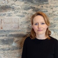 Photograph of Dr Rosie Lavan, with shoulder-length reddish-blonde hair, standing in front of a textured stone wall. She is wearing a simple black top and looking calmly at the camera.