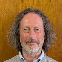 Headshot of a Dr Patrick Roycroft, wearing a light gray sweater over a blue collared shirt, against a wooden background.