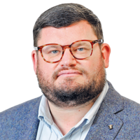 Portrait of Donal Fallon wearing tortoiseshell glasses, a blue tweed blazer and a light blue shirt, against a plain background.