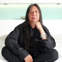 Portrait of John Rocha with his long dark hair loose, sits cross-legged on a white bed, wearing black clothing and silver jewellery. He rests his chin on one hand and looks directly at the camera, with a pale green and white headboard behind him.