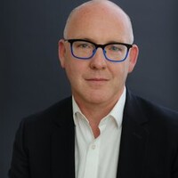 Portrait of a Kevin Rafter wearing blue-framed glasses, a black suit jacket and a white shirt, looking directly at the camera against a dark grey background.