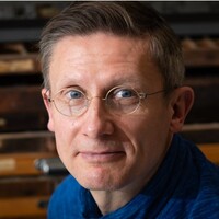 Portrait of Marco Sonzogni wearing round glasses and a blue scarf, looking towards the camera against a dark wooden background.