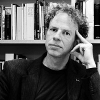 Black-and-white portrait of Matthew Hollis seated in front of bookshelves, resting his head against one hand and looking towards the camera.